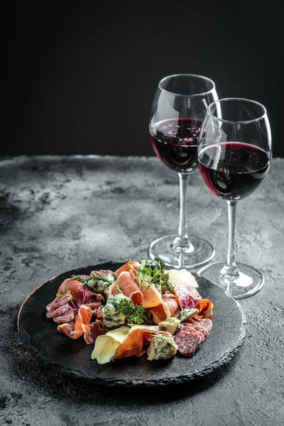 Antipasto platter. Glass of red wine with various cheeses and prosciutto on a black background. vertical image, place for text. Food recipe background. Close up.