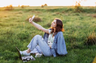 Kadın cep telefonuyla görüntülü konuşma yapıyor. Yeşil çimlerin üzerinde oturan gülümseyen kız bilgisayar ve cep telefonu kullanıyor. Uzaklık öğrenme çevrimiçi eğitim ve çalışma,