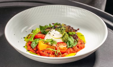İtalyan burrata peyniri, domates konçertosu, pesto sosu ve beyaz tabakta taze fesleğen ile servis edilir..