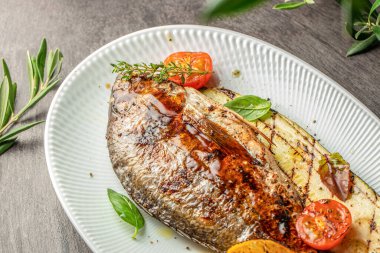 Izgara dorado balığı, kabak, kiraz domatesleri ve fesleğen yapraklarıyla servis edilir, üzerine zeytin yağı ve beyaz bir tabakta otlar serpilmiştir..