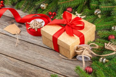 Köknar dalları, takılar, sicimler ve kırsal ahşap arka planda tatil süslemeleriyle çevrili kırmızı kurdeleyle sarılı Noel hediyesi kutusu.