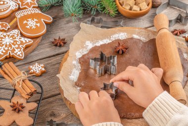 Noel kurabiyelerini kağıt üzerinde metal kesicilerle kesen eller, merdane, baharat, dekore edilmiş kurabiyeler ve köknar dalları ile şenlikli pişirme süreci, masa üzerindeki üst görünüm..