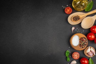 Fresh cooking ingredients on dark stone background with tomatoes, basil, garlic, spices, olive oil and wooden spoons arranged as a flat lay copy space recipe or food design projects.