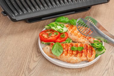 Top view of grilled chicken breast served on a white plate with fresh tomato slices, cucumber, basil, and green onions. Juicy roasted meat with grill marks placed beside an electric grill