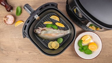 Top-view image of a whole fresh fish placed inside an air fryer basket with lemon slices and basil leaves on a wooden table.