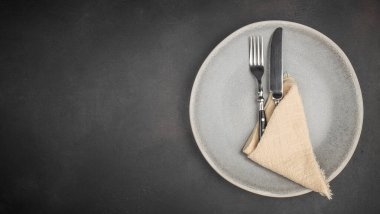 Top view of an elegant table setting featuring a ceramic plate with a fork and knife placed on a folded beige napkin over a dark textured background. Minimalist composition