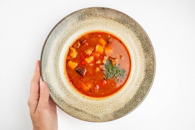 Kırsal bir kasede servis edilen sebzeli ve etli geleneksel sıcak kırmızı çorba, temiz beyaz arka planda, üst tarafta..