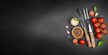 Minimum yemek pişirme arka planı, kiraz domatesleri, fesleğen, sarımsak, baharat, mutfak bıçağı ve çatal ile siyah tahta yüzeyde büyük bir kopya alanı, yemek tarifi veya reklam tasarımı için..