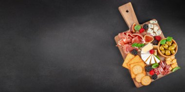 Pişmiş etler, çeşitli peynirler, zeytinler, krakerler, incirler ve böğürtlenlerle dolu zarif bir antipasto tahtasının en üst görüntüsü. Koyu bir arkaplanda servis panosunun üzerinde düzenlenmiş, metin için fotokopi alanı ile.