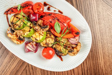 Kabak, kırmızı dolma biber, mantar, soğan ve kiraz domatesleri dahil olmak üzere ızgara mevsimlik sebzelerin üst görüntüsü, ahşap masa üzerinde beyaz tabakta servis ediliyor.