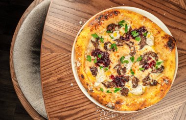 Üstünde et, erimiş peynir, karamelize soğan reçeli ve ahşap arka planda beyaz tabakta taze otlar bulunan zanaatkar pizzanın üst görüntüsü.