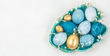 Zarif mavi, turkuaz ve altın sarısı Paskalya yumurtaları seramik kasede küçük çiçeklerle süslenmiş, üst görünüm düz beyaz zemin üzerinde, metin için büyük kopya alanı var.