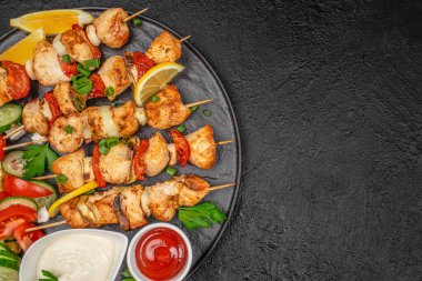 Grilled chicken skewers with tomatoes, onions and herbs served with creamy and tomato sauces on black plate over dark textured background, top view
