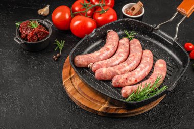 Izgara tavasında çiğ sosis, biberiye, domates, baharat ve sosla birlikte koyu taş arka planda, barbekü, menü ve yemek reklamları için kırsal pişirme süreci konsepti.