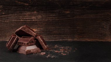 Siyah yüzeye yığılmış siyah çikolata parçaları, kaba ahşap arka plan, tatlı reklamları için kopyalama alanı, gıda markası ve çikolata tasarımı.