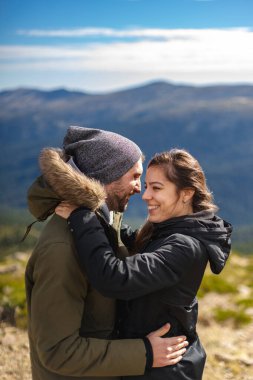 Aşık genç bir çift ve kışlık giysiler, güneşli bir günde dağ sırasının önünde sarılıp gülümsüyor.