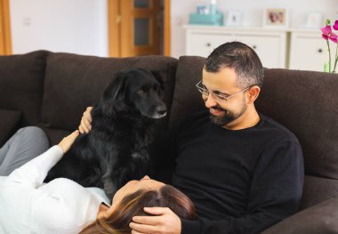 Evdeki kanepede siyah köpekleriyle birlikte mutlu genç bir çift. Kadın kafasını partnerinin bacaklarına yaslıyor..