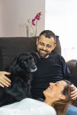 Evdeki kanepede siyah köpekleriyle birlikte mutlu genç bir çift. Kadın kafasını partnerinin bacaklarına yaslıyor..