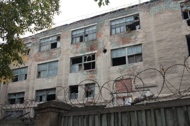 Çitin arkasındaki ön cephesinde kırık camlar olan terk edilmiş bir bina.