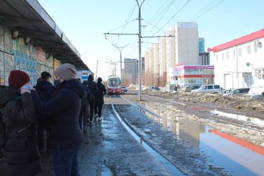 Russia, Novosibirsk 04.04.2021: masked passengers wait for a tram on rails in a puddle for a ride in a dirty city in the spring in the slums