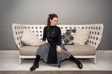 Gri odadaki bir kanepede oturan genç kadın manken. Siyah ve gri elbiseli güzel esmer kadın içeride poz veriyor.