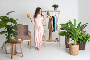 Pembe elbiseli vejetaryen kıvırcık kadın elinde ananas ve portakal tutuyor. Kahvaltıda taze meyve seçiyorum.