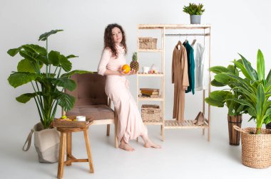 Pembe elbiseli vejetaryen kıvırcık kadın elinde ananas ve portakal tutuyor. Kahvaltıda taze meyve seçiyorum.