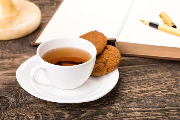 Fildişi çay bardağı tatlı kurabiye, kalem ve kitap ahşap palet üzerinde