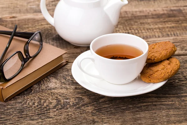 Taza de té de marfil con galletas dulces, vasos y libro en madera pálida — Foto de Stock