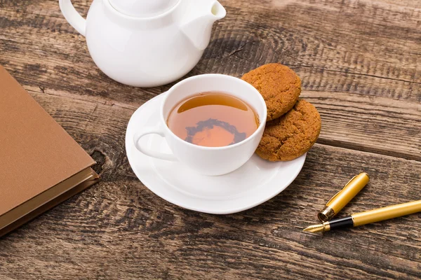 Taza de té de marfil con galletas dulces, pluma y libro en paleta de madera — Foto de Stock