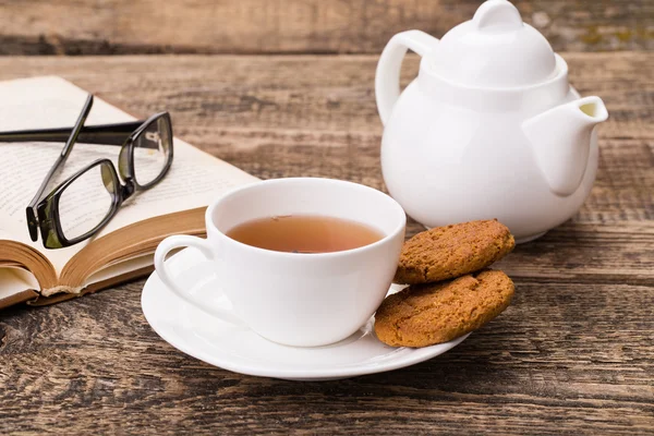 Tazza di tè avorio con dolce biscotto, bicchieri e libro su legno pallido — Foto Stock