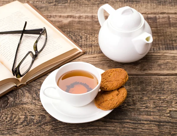 Xícara de chá de marfim com biscoito doce, óculos e livro em madeira pálida — Fotografia de Stock