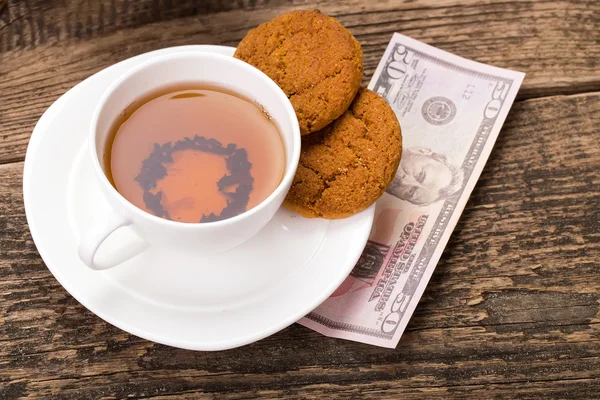 Taza de té de marfil con galletas dulces, y dinero en dólares de madera pálida — Foto de Stock