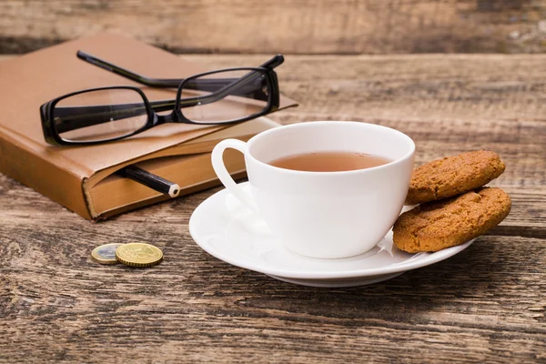 Taza de té de marfil con galletas dulces, vasos, dinero y libro sobre madera — Foto de Stock