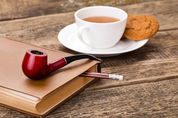 Xícara de chá de marfim com biscoito doce, cachimbo de tabaco e livro em madeira — Fotografia de Stock