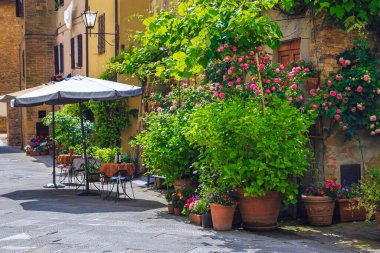 Eski asfalt sokakta masaları ve sandalyeleri olan harika küçük bir sokak kafesi. Pienza, Toskana, İtalya ve Avrupa 'da renkli çiçeklerle süslü bitkiler ve dekore edilmiş duvarlar.