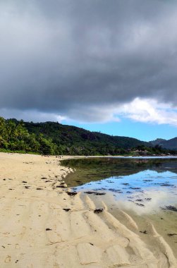 Tropical beach in the Seychelles Islands featuring white sand, clear shallow water, lush palm trees, and a moody sky