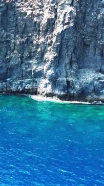 Derin mavi okyanusu çevreleyen engebeli bir volkanik kayalığın havadan görünüşü. Kristal berrak turkuaz su kayalık kıyıya hafifçe sıçrıyor.