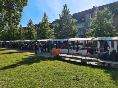 Neumuenster, Almanya - 28. Eylül 2025: Güneşli bir günde bir parkta ikinci el giysi ve eşya alışverişi yapan bir çok insanla açık hava bit pazarı tezgahları