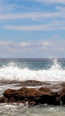 Dinamik okyanus dalgaları engebeli kıyı kayalarına çarpar, açık mavi gökyüzüne köpük püskürterek yumuşak bulutlar yayar, ham tropikal enerjiyi ve saf deniz manzarası enerjisini yakalar.