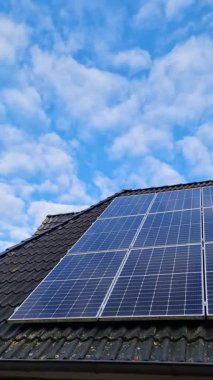 Bulutlu mavi gökyüzü arka planına sahip bir evin çatısındaki güneş panelleri yenilenebilir enerji sağlıyor