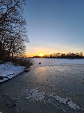 Donmuş göl ılık günbatımı renklerini yansıtıyor soğuk bir kış akşamında karla kaplı kıyılarda.