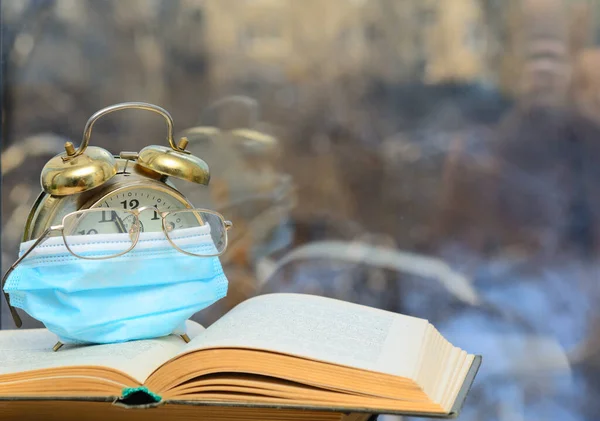 Vintage alarm clock partially covered by a medical mask on a white background. Nearby lies a red ball - a New Year\'s gift. Concept - a pandemic during the New Year