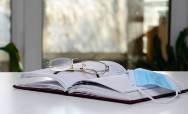 Açık günlükte gözlük ve tıbbi maske var. Pencereden ışık düşüyor. Yalıtım konsepti, yeni bir günün planlanması.