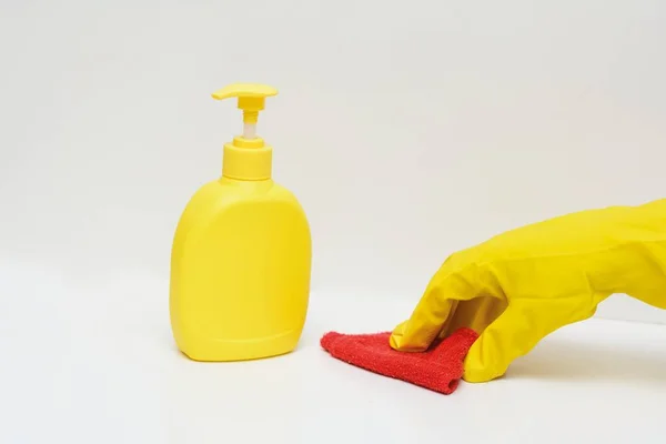 On a white background, a yellow bottle with liquid soap. A hand in a yellow glove rubs the surface of the table with a red rag. Concept - hygiene, cleanliness