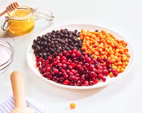 A horizontal composition with frozen sea buckthorn, cranberry and blueberry berries on a white plate, a jar of honey and sugar on white background. Fermentation of berries. Home made berries mors, jam