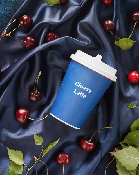 Cherry latte, new kind of coffee. Food composition with craft cup of coffee, cherries and leaves on a blue, satin, folded background. Coffee to go. Tasty, summer drinks concept. Flat lay