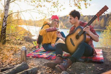 Bir çift, sonbahar ağaçları ve nehrin arka planına karşı doğada dinleniyor. Adam gitar çalıyor.