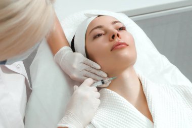 Doktor kozmetik uzmanı, güzellik salonundaki genç ve güzel bir kadının cildindeki kırışıklıkları sıkmak ve düzleştirmek için canlandırıcı yüz enjeksiyonu prosedürünü uyguluyor..
