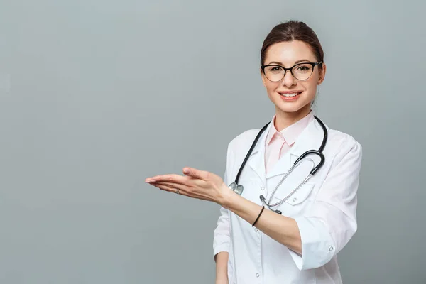 Vriendelijk lachende jonge vrouwelijke arts. Hand wijzend naar kopieerruimte. Geïsoleerd op een grijze achtergrond. — Stockfoto